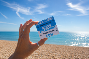 Hand holding Peppersmith gum and mints against a sunny beach and sea backdrop.
