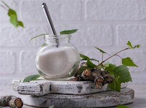 A white brick background with plants and wood. On the wood there is a glass jar with Xylitol in and spoon. 