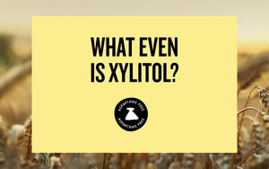 Yellow background with bold black text reading ‘What even is xylitol?’ and a small circular badge underneath that says ‘Aspartame Free’, set over a blurred wheat field background.